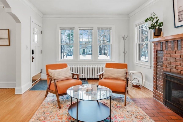 Une pièce avec deux fauteuils orange, une table basse en verre, une cheminée et une vue sur l’extérieur grâce à de grandes fenêtres.