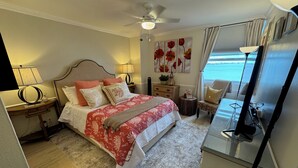 Ein Schlafzimmer mit einem Bett, Nachttischen, einer Lampe, einem kleinen Tisch mit einer Pflanze und einem Fenster mit Vorhängen.