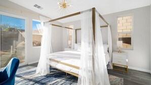 A modern bedroom with a four-poster bed, a large window, a blue chair, and a nightstand with a lamp.