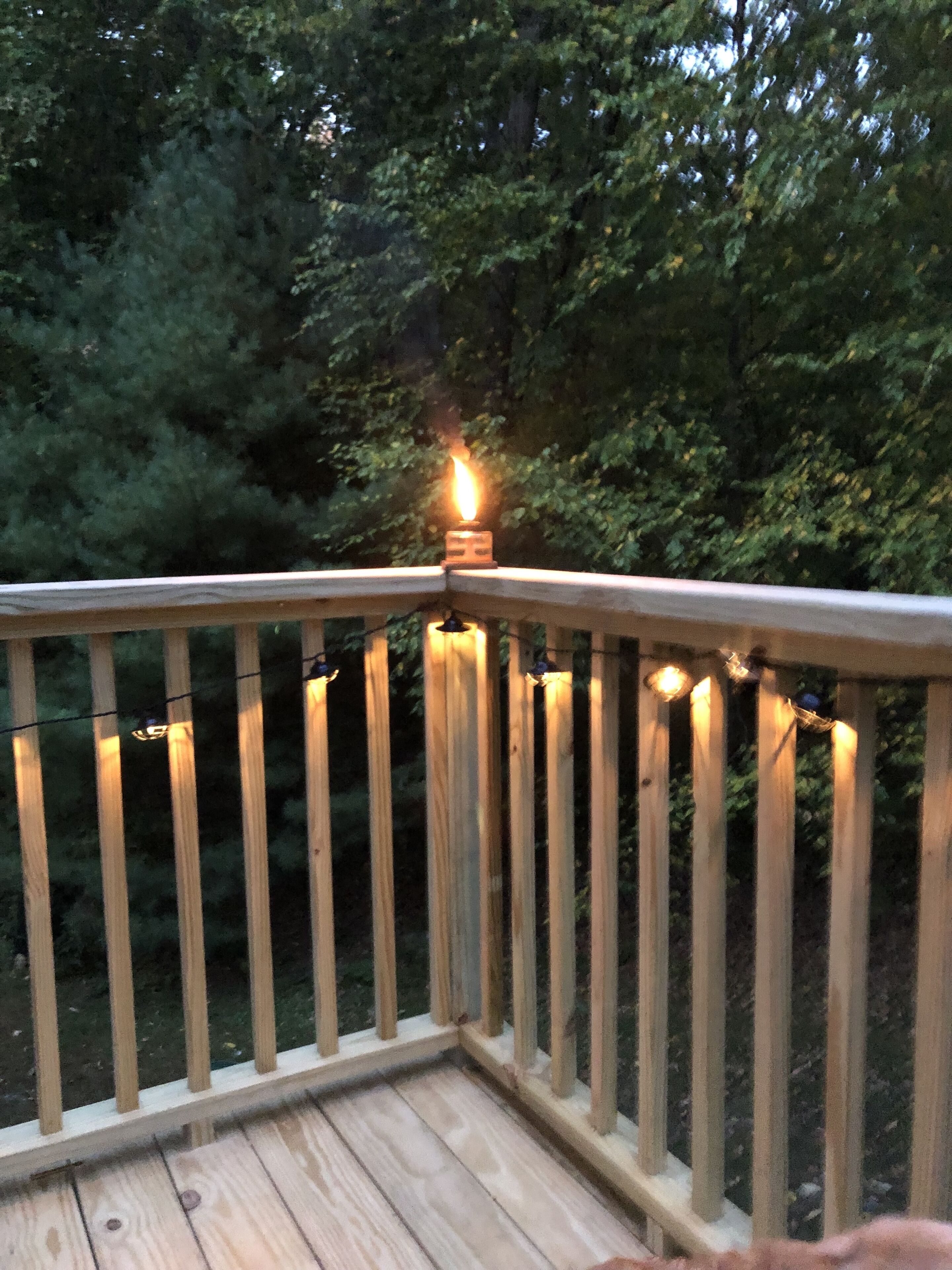 Terraza de madera con barandilla, luces colgantes y una vela encendida.