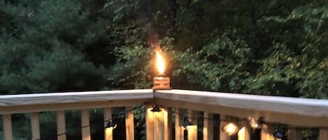 Terraza de madera con barandilla, luces colgantes y una vela encendida.