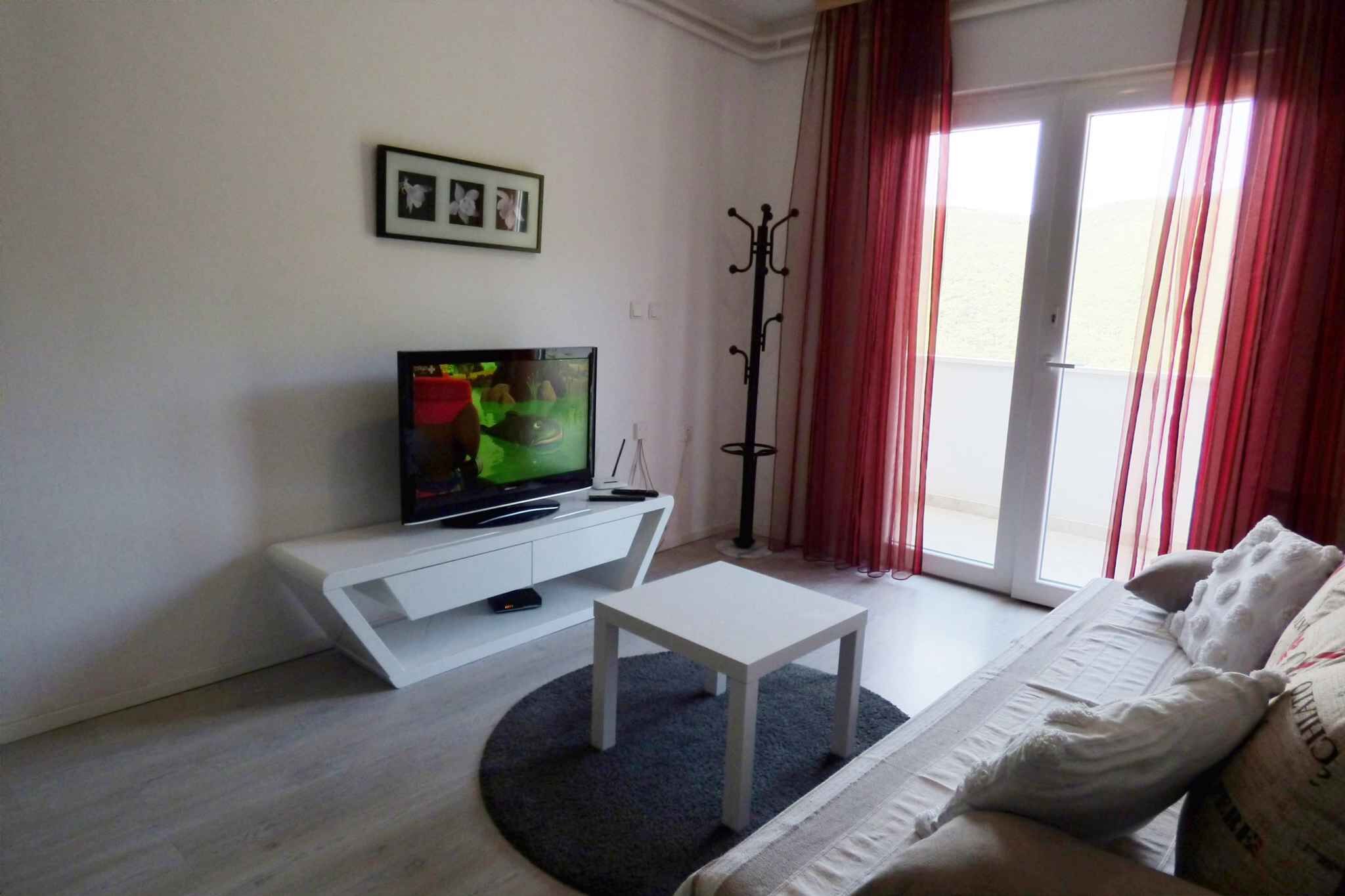 Ein modernes Wohnzimmer mit einem Flachbildfernseher auf einer weißen Konsole, einem weißen Sofa und einem großen Fenster mit roten Vorhängen.