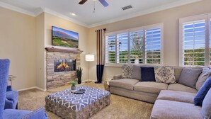A living room with a stone fireplace, a ceiling fan, and a large window with blinds.