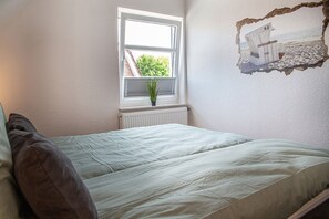 A bedroom with a bed, a window with blinds, a radiator, and a wall-mounted picture. - 50136 "Deichkajüte 5" - Modern, fresh and high-quality: this terraced end-terrace house (Wittmund)
