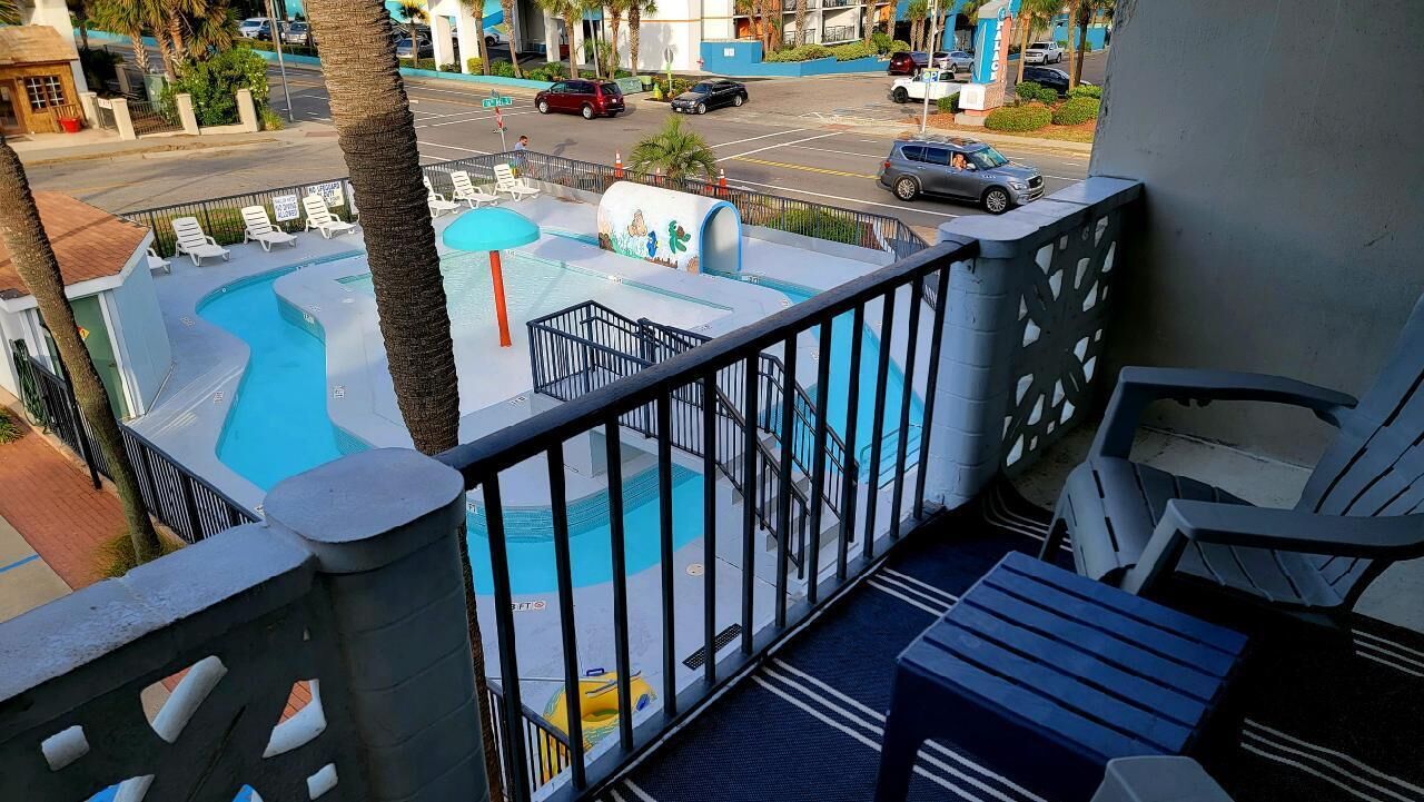 A balcony with a view of a swimming pool, lounge chairs, and a street view.