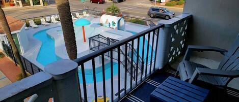 A balcony with a view of a swimming pool, lounge chairs, and a street view.