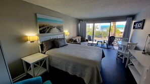Quarto de hotel com cama, escrivaninha, cadeira, varanda com mesa e cadeiras, e uma pintura representando uma cena de praia.