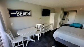 Quarto de hotel com cama, escrivaninha com cadeira, TV fixada na parede e placa indicando a direção para a praia.