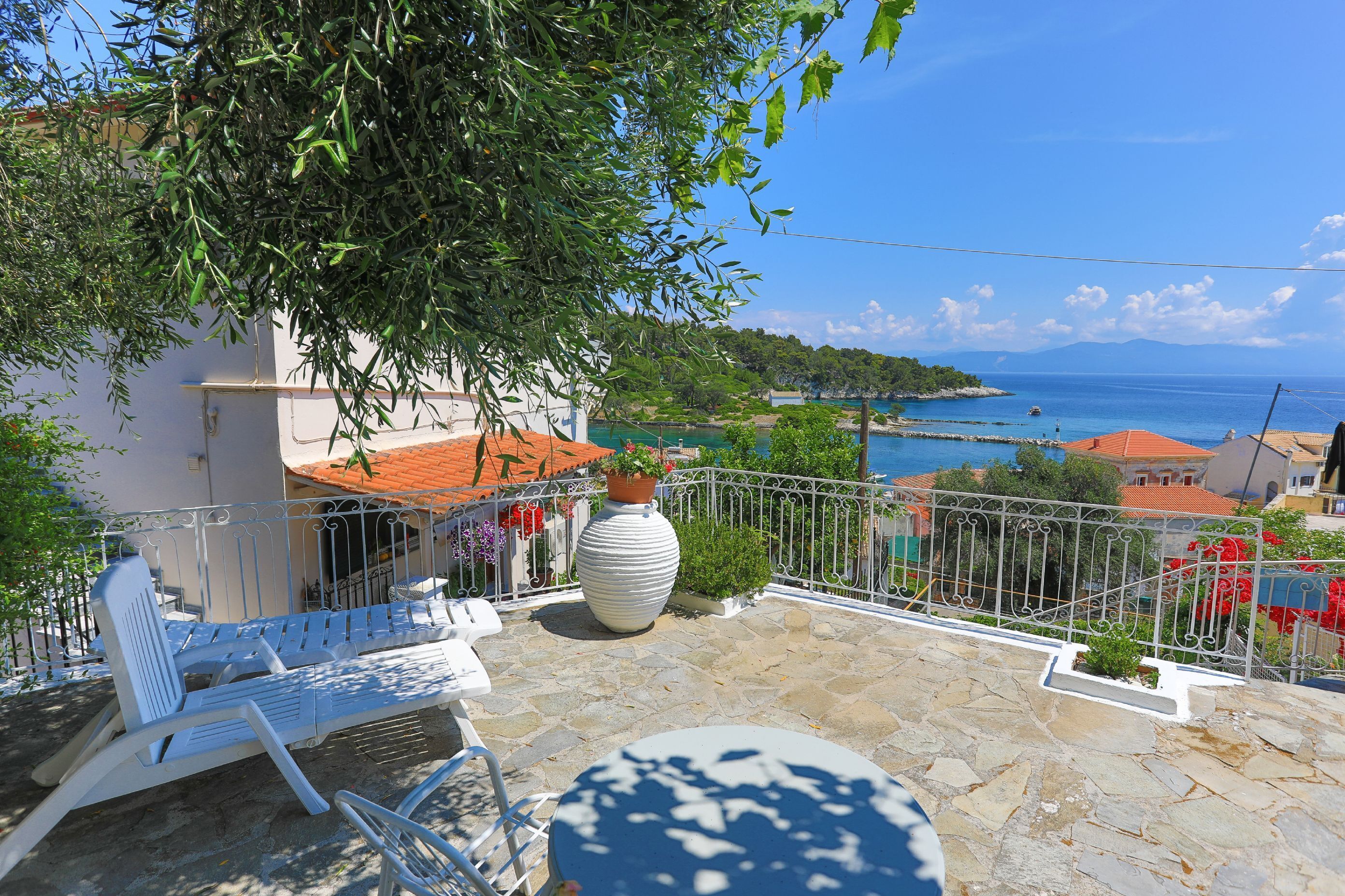 A terrace with white lounge chairs, a large potted plant, and a view of the sea and buildings.