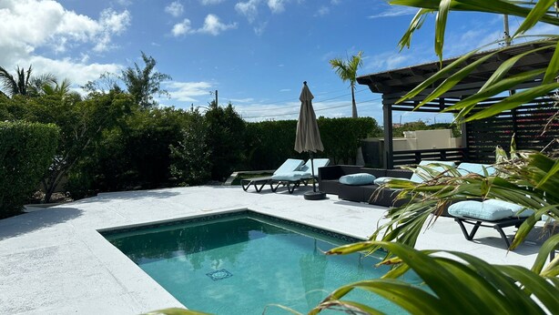 A rectangular swimming pool with a tiled edge, surrounded by a concrete deck. There are lounge chairs and a covered seating area with an umbrella. - Beautiful villa West with pool- 5 minutes to Gracebay beach (Providenciales)