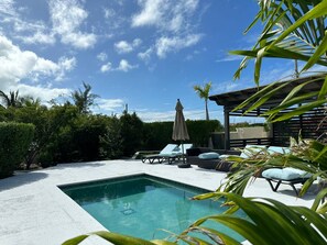 Ein rechteckiger Pool mit einer gefliesten Kante, umgeben von einer Betonplattform. Liegestühle und eine überdachte Sitzecke mit Sonnenschirm sind vorhanden.