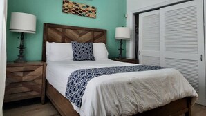 A bedroom with a wooden headboard, a bed with a patterned bedspread, two bedside lamps, a wall-mounted air conditioner, and a closet with louvered doors. - Beautiful villa West with pool- 5 minutes to Gracebay beach (Providenciales)
