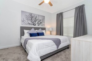 Un dormitorio con cama, cabecera, mesita de noche, lámpara, ventana con cortinas, ventilador de techo y una obra de arte colgada en la pared.