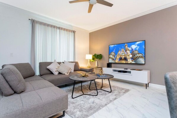 Una sala de estar moderna con un sofá gris, una mesa de centro, un televisor mostrando un edificio y un ventilador de techo.
