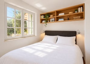 Une chambre avec une grande fenêtre, un lit recouvert de linge de maison blanc, une tête de lit en bois et une étagère avec des livres et des objets décoratifs.