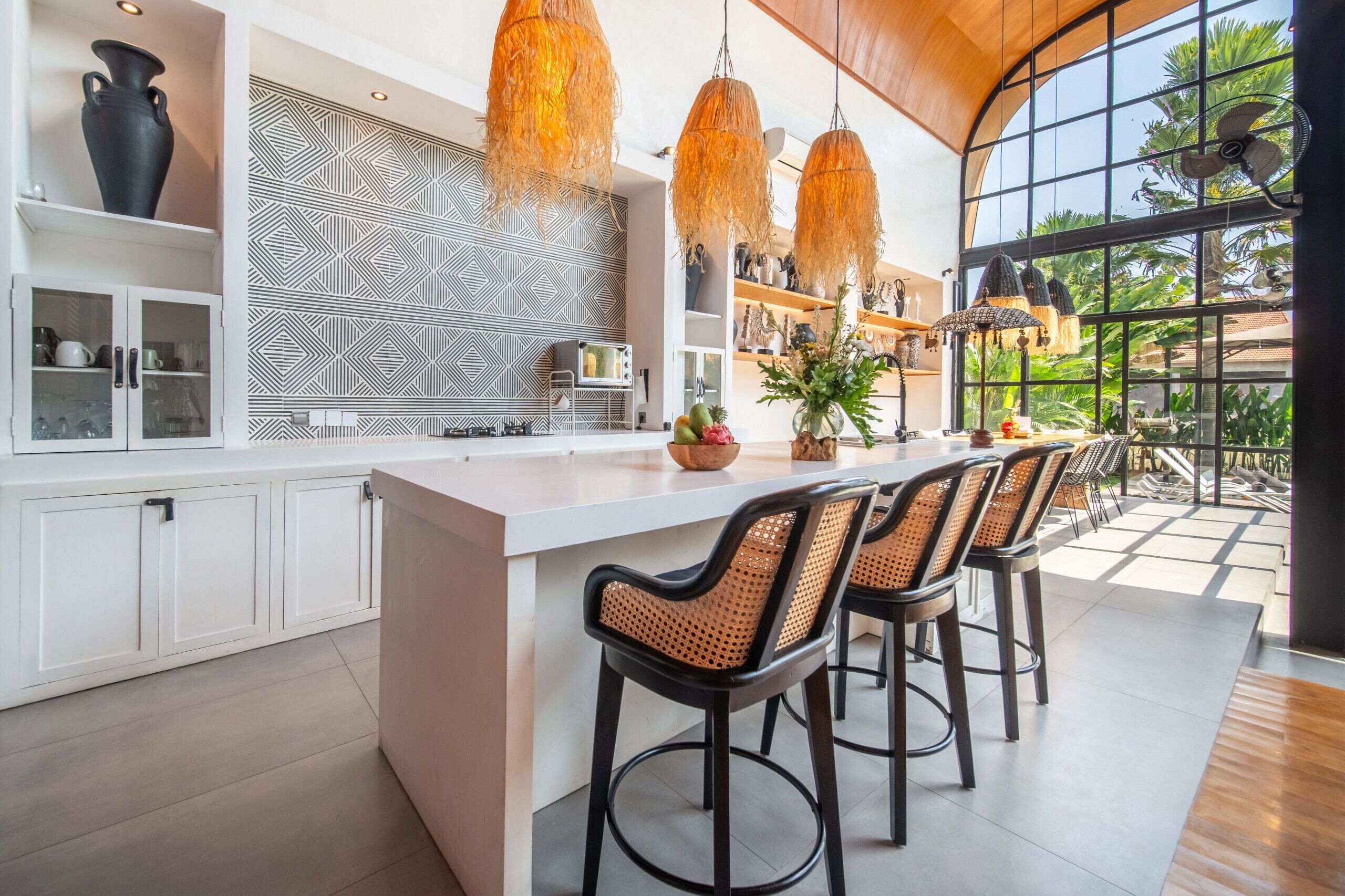 Cocina moderna con una isla grande, muebles blancos y luces colgantes naranjas.