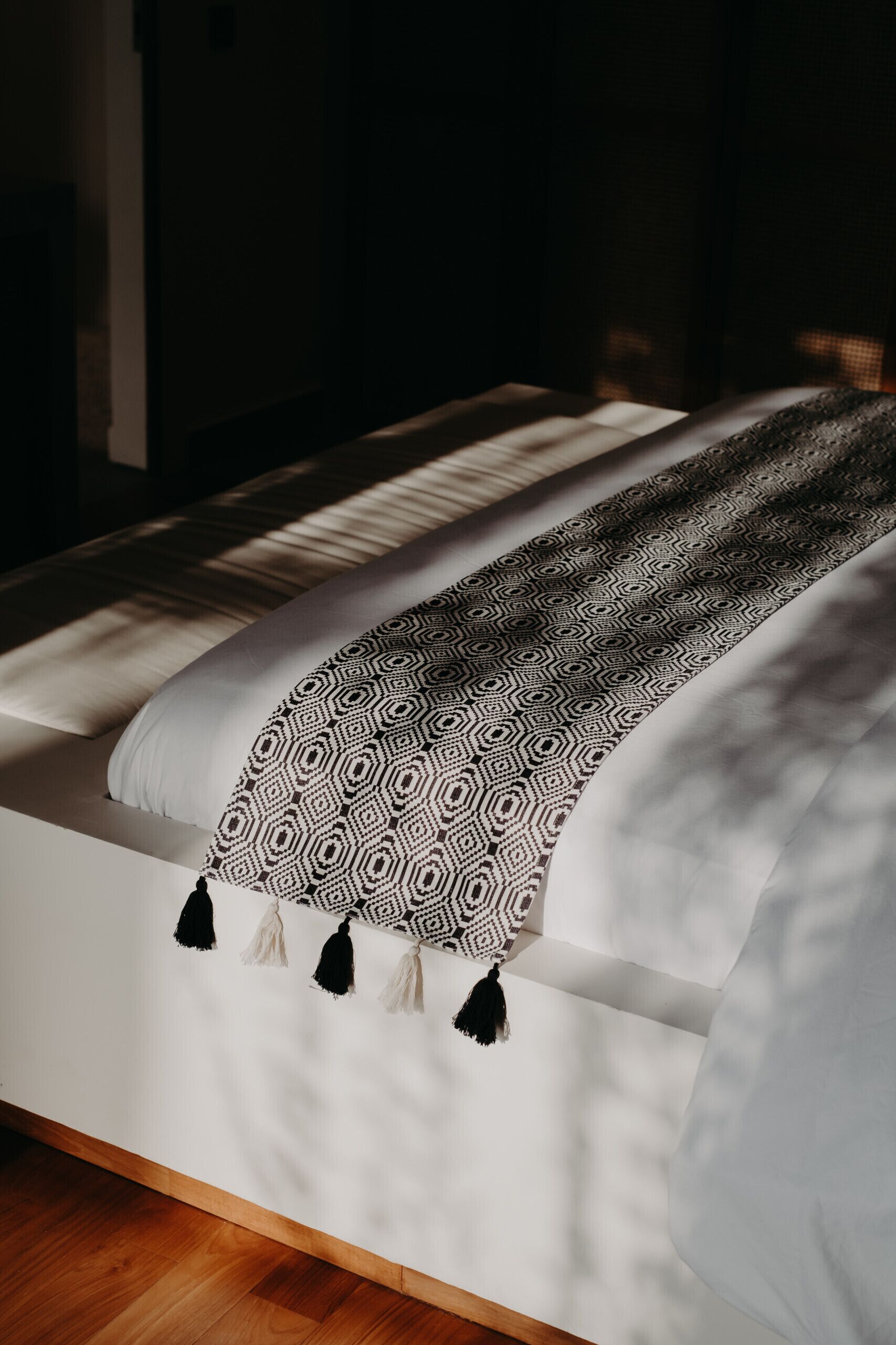 A bed with a patterned blanket and tassels, a wooden floor, and a white wall.