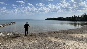 A person standing on a pebbly beach with a calm sea and a forested island in the distance.