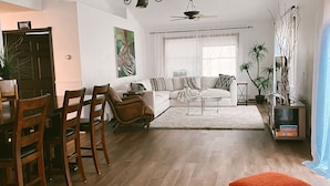 A spacious living area with a dining table, chairs, a sofa, and a coffee table. There are ceiling fans, a chandelier, and a large window with blinds.