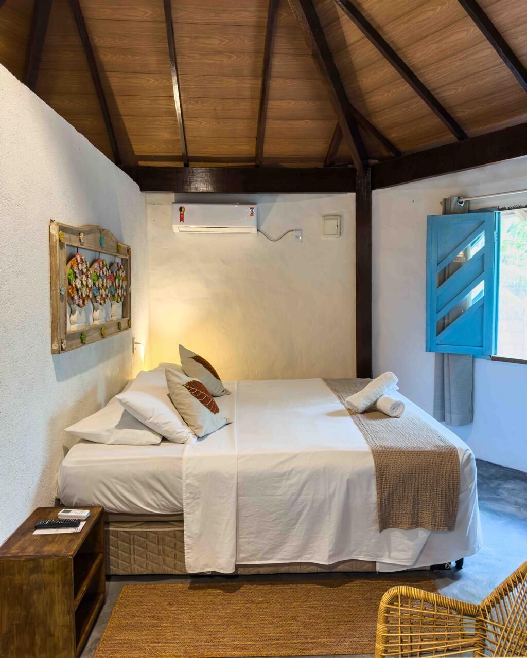A bedroom with a wooden ceiling, a bed with white bedding, a small bedside table, a wicker chair, and a window with a view.