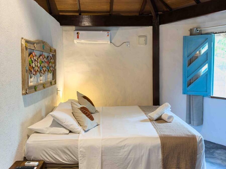 A bedroom with a wooden ceiling, a bed with white bedding, a small bedside table, a wicker chair, and a window with a view.
