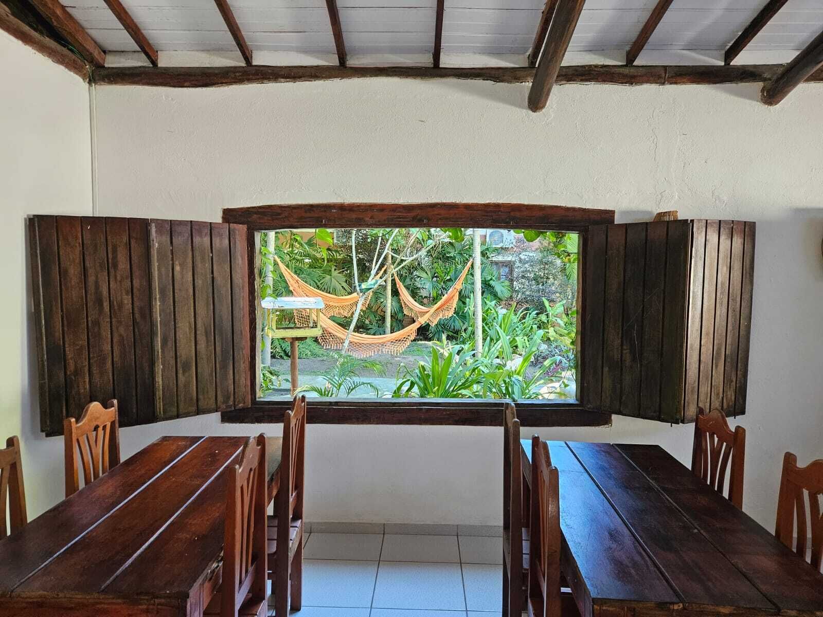 A dining area with a large window overlooking a garden, featuring wooden furniture and a hammock.