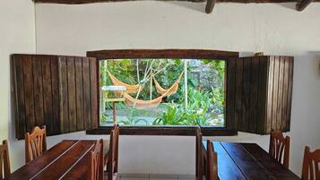A dining area with a large window overlooking a garden, featuring wooden furniture and a hammock.