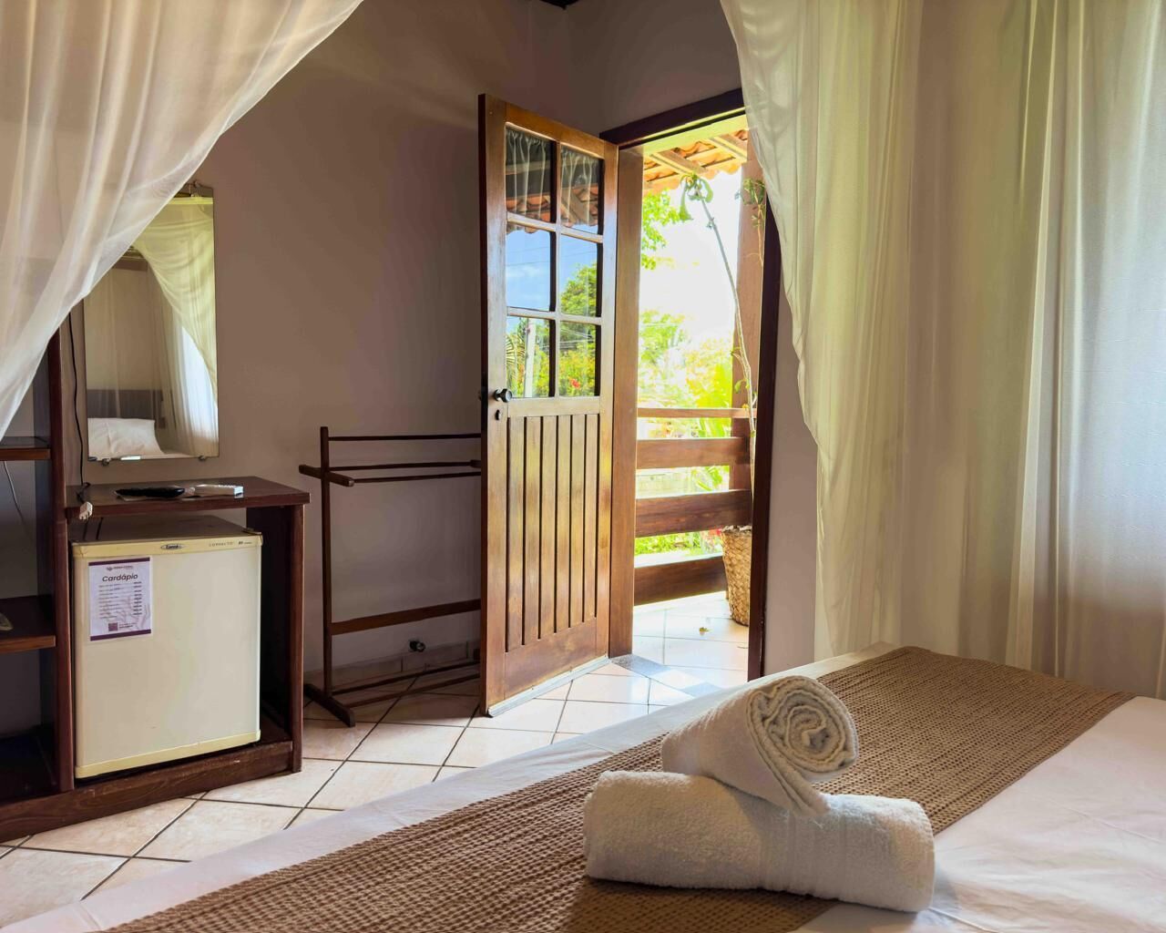 A bedroom with a bed, a desk, a small refrigerator, and a wooden door leading to a balcony.
