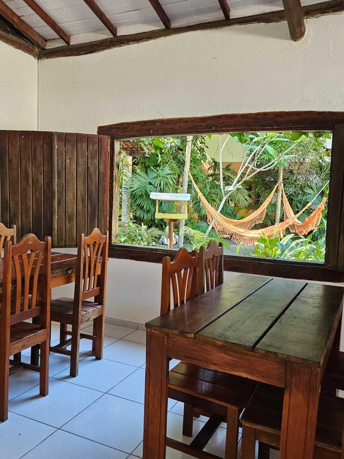Uma sala de jantar com mesa e cadeiras de madeira, uma janela grande com vista para uma rede e vegetação.