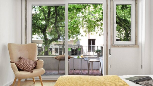 Una habitación con cama, una silla, una mesita con una planta y vistas a árboles a través de una puerta corredera.