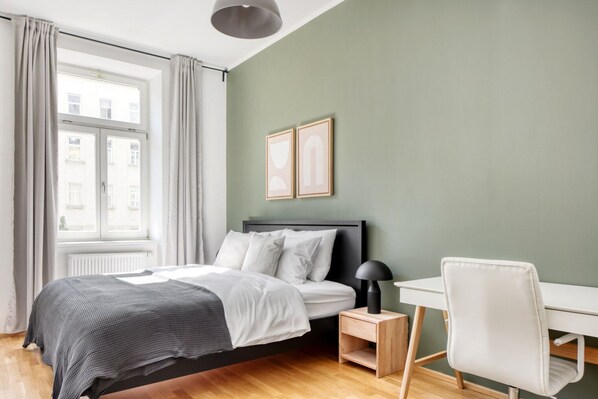Une chambre à coucher comprenant un lit, une table de chevet, une lampe, une chaise et une fenêtre avec des rideaux.