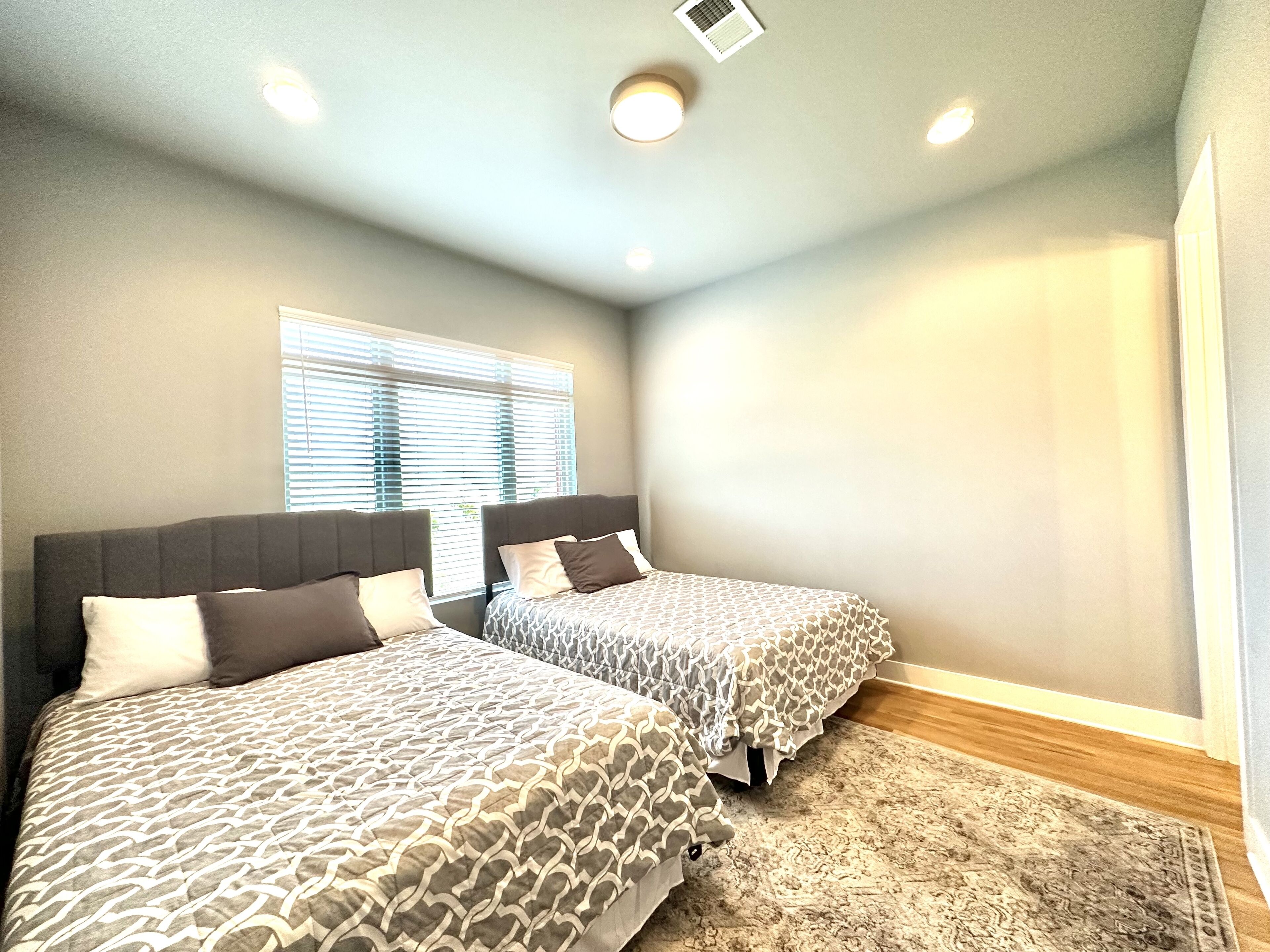 A bedroom with two beds, a window with blinds, a ceiling light, and a patterned rug.