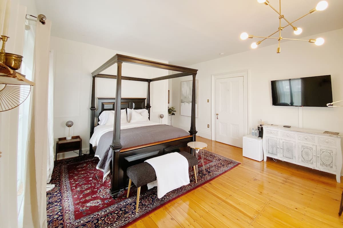 A four-poster bed with a dark finish, a white bedside table, a small stool, a red patterned rug, and a modern chandelier.