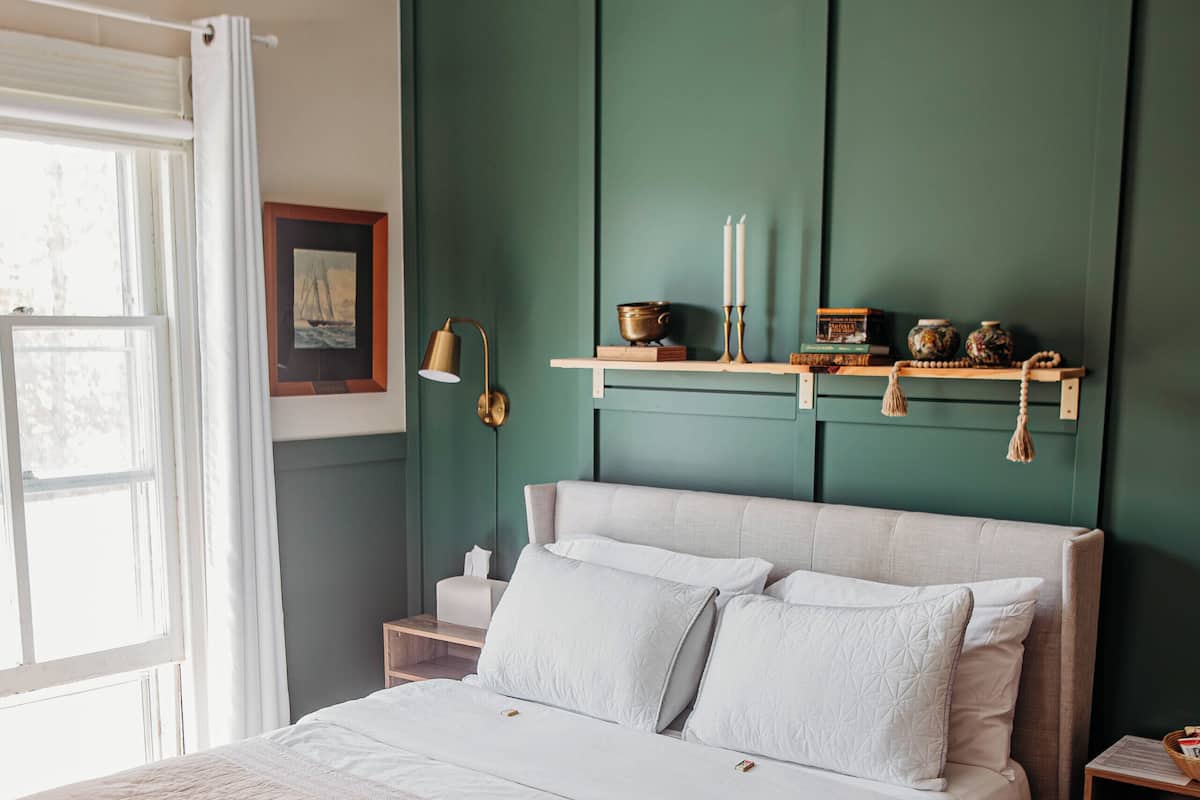 A neatly arranged bedroom with a large bed, a bedside table, a nightstand, and a wall-mounted shelf with decorative items.