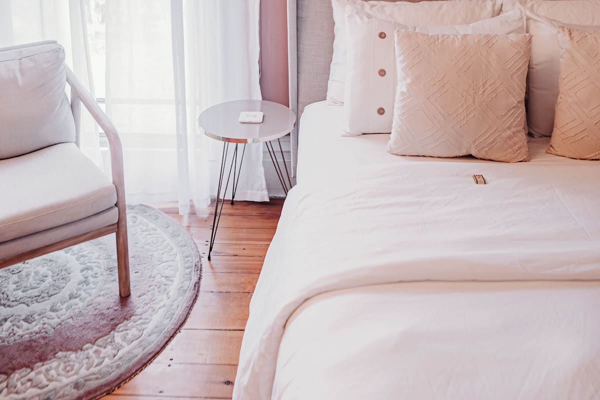 A bedroom with a bed, a chair, a small table, and a window with sheer curtains.