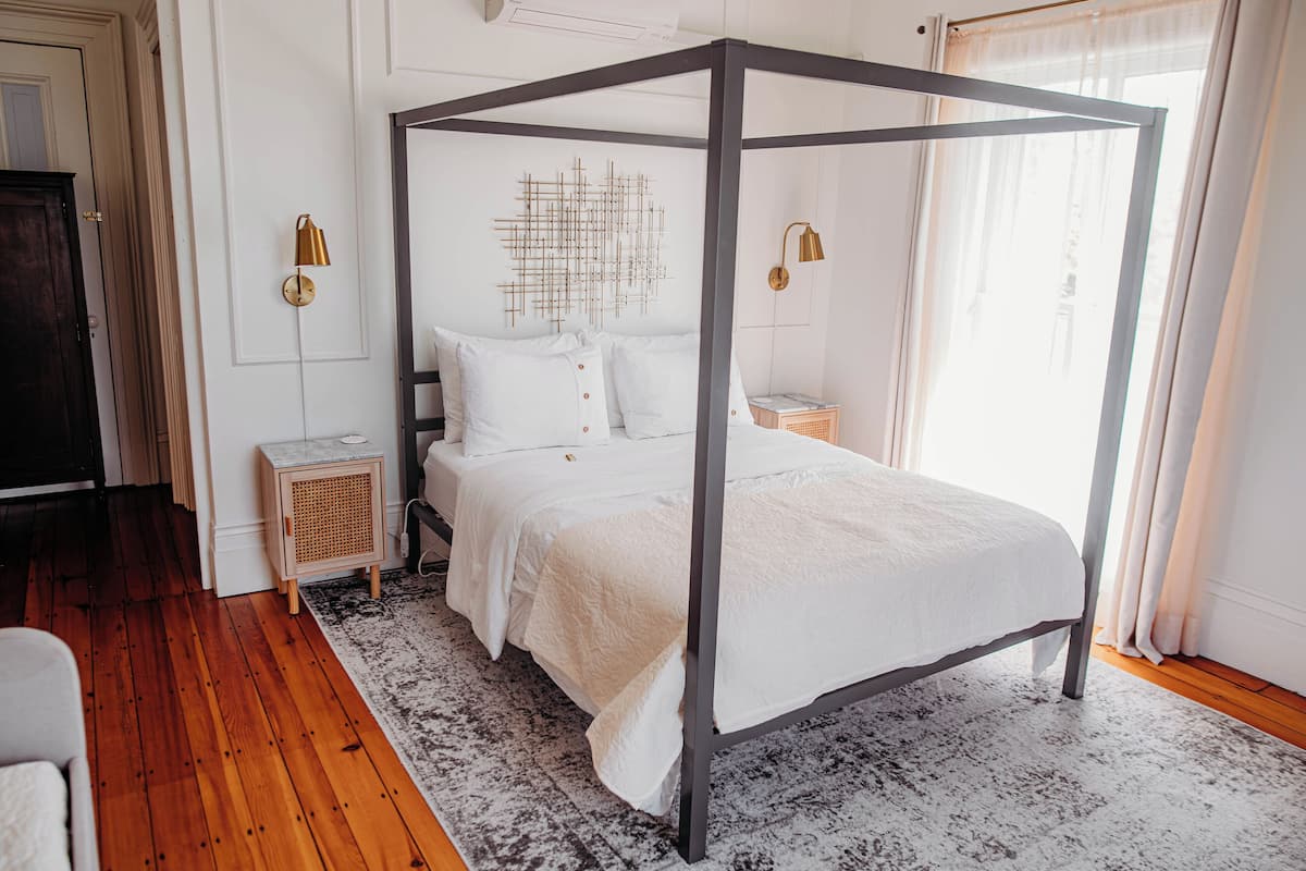 A four-poster bed with white bedding, a rattan nightstand, a wall-mounted lamp, and a geometric wall art piece.