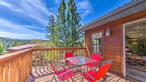 A wooden deck with a glass table and red chairs, overlooking a forested area.