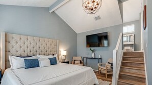 A bedroom with a tufted headboard, a large bed with white bedding, a chandelier, a television mounted on the wall, and a wooden staircase. - Lakeside Paradise, lake views, hot tub (Canyon Lake)