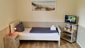 Ett litet, ombonat rum med en säng, ett nattduksbord med en lampa, en tv på ett stativ och en hylla med böcker.