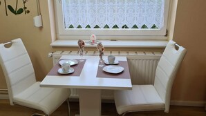 En matplats med ett vitt bord och två vita stolar med vadderade sitsar. Ett fönster med spetsgardiner sitter ovanför bordet, och en element är placerad under fönstret.