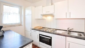 A modern kitchen with white cabinets, a black countertop, a built-in oven, a dishwasher, and a window with blinds. - Haushälfte "kuddel" 104 (St. Peter-Ording)