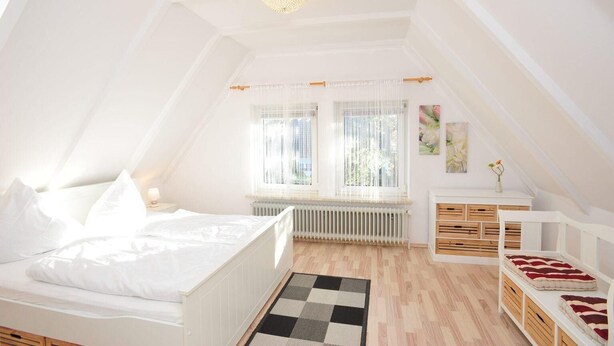 A white, sloped-roof bedroom with a bed, a radiator, a window with sheer curtains, and a small bedside table with a lamp. - Haushälfte "kuddel" 104 (St. Peter-Ording)