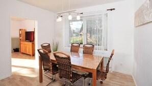 A dining area with a wooden table and wicker chairs, a television in the background, and a view through sheer curtains. - Haushälfte "kuddel" 104 (St. Peter-Ording)