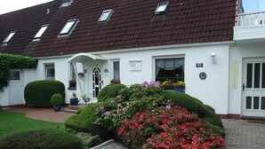 A white building with a red-tiled roof, a small garden with red flowers, and a neatly trimmed hedge. - App. Liebrecht OG Links (St. Peter-Ording)