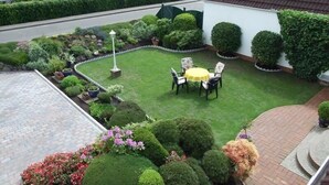 A well-maintained garden with a lawn, a round table with chairs, and various plants and shrubs. - App. Liebrecht OG Links (St. Peter-Ording)
