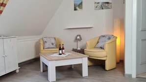 A cozy room with a white cabinet, two yellow armchairs with star-patterned pillows, a small wooden table with a bottle and a plate, and a lamp on a side table. - App. Liebrecht OG Links (St. Peter-Ording)