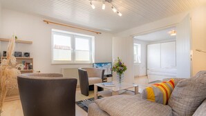 A modern living room with a sofa, armchairs, a coffee table, and a TV. There are windows with blinds, a radiator, and a bookshelf with books and decorative items. - Wohnung A (St. Peter-Ording)