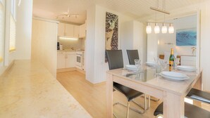 A modern kitchen with white cabinets, a dining area with a glass table, and a view of the sea through a window. - Wohnung A (St. Peter-Ording)