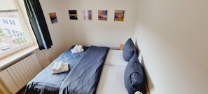Uma cama de solteiro com edredom e almofadas azuis listradas, uma janela com vista para edifícios e quatro quadros na parede.