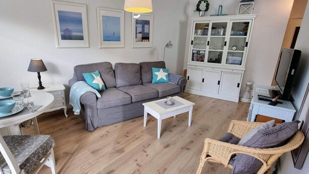 Ein Wohnzimmer mit grauem Sofa, einem weißen Schrank, einem Korbstuhl und einem Esstisch mit blauer Schüssel und Gläsern.